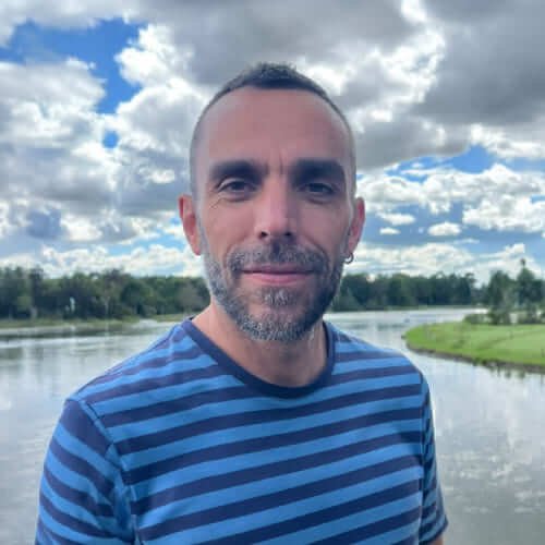 Retrato de hombre con camiseta a rayas azules frente a un río y cielo nublado.