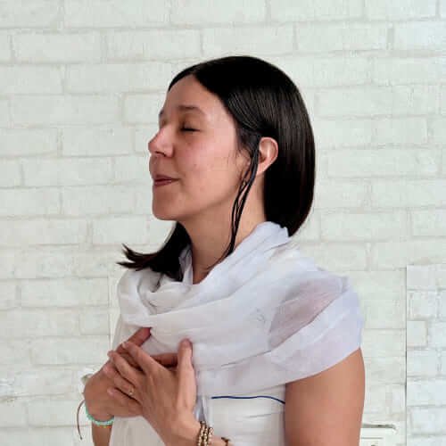 Mujer con los ojos cerrados meditando con las manos en el pecho frente a pared blanca.