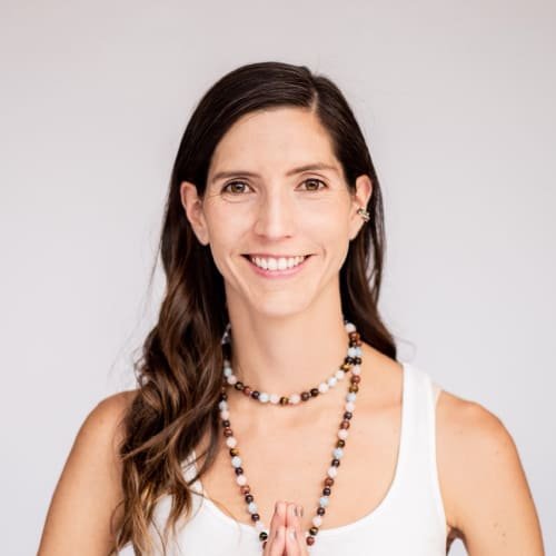 Mujer sonriente con collar de cuentas y manos juntas en gesto de gratitud sobre fondo blanco.