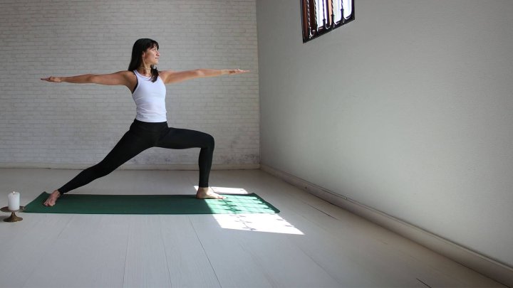 Mujer realizando postura de yoga guerrero II en tapete verde, con velas y luz natural, Yogatón apoya a UNICEF para cada infancia.